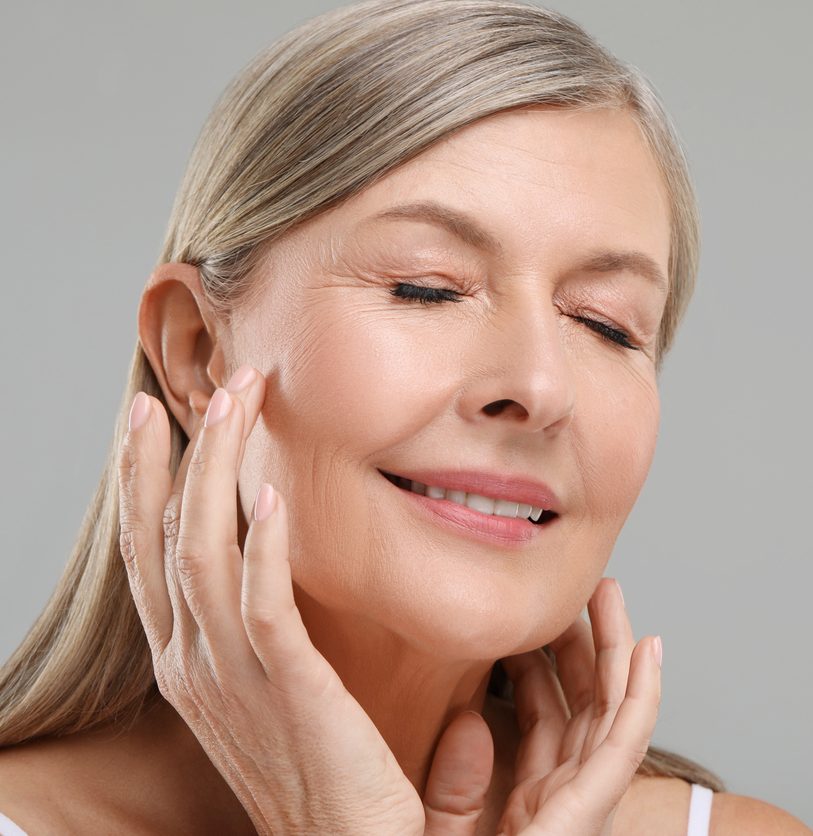 Smiling mature woman with closed eyes touching her face against a gray background