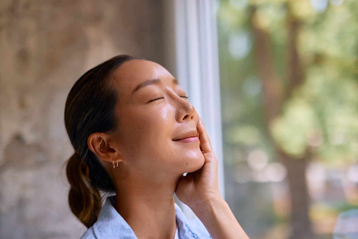 Relaxed woman touching her face by a window, eyes closed, enjoying sunlight